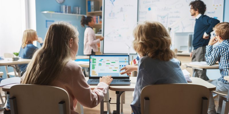 Elementary School Science Class: Over the Shoulder Little Boy and Girl Use Laptop with Screen Showing Programming Software. Physics Teacher Explains Lesson to a Diverse Class full of Smart Kids